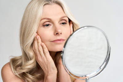 Closeup portrait of gorgeous happy middle age woman looking at mirror touching her skin enjoying treatment for dry skin. Advertising of antiaging beauty skin care products.